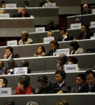 Participants sit in rows at a WTO ministerial meeting, listening.